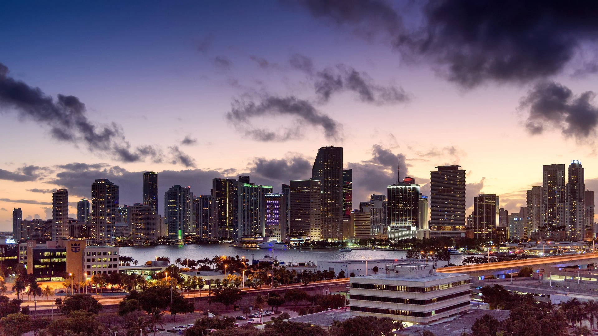 A skyline of Miami at dusk with lit skyscrapers reflecting on the water, silhouetted clouds, and a purple and orange sunset